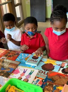 Three children in masks select a book from a spread of books depicting Black characters