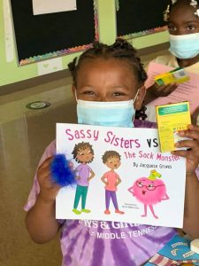 Girl in mask smiles while holding book featuring Black characters