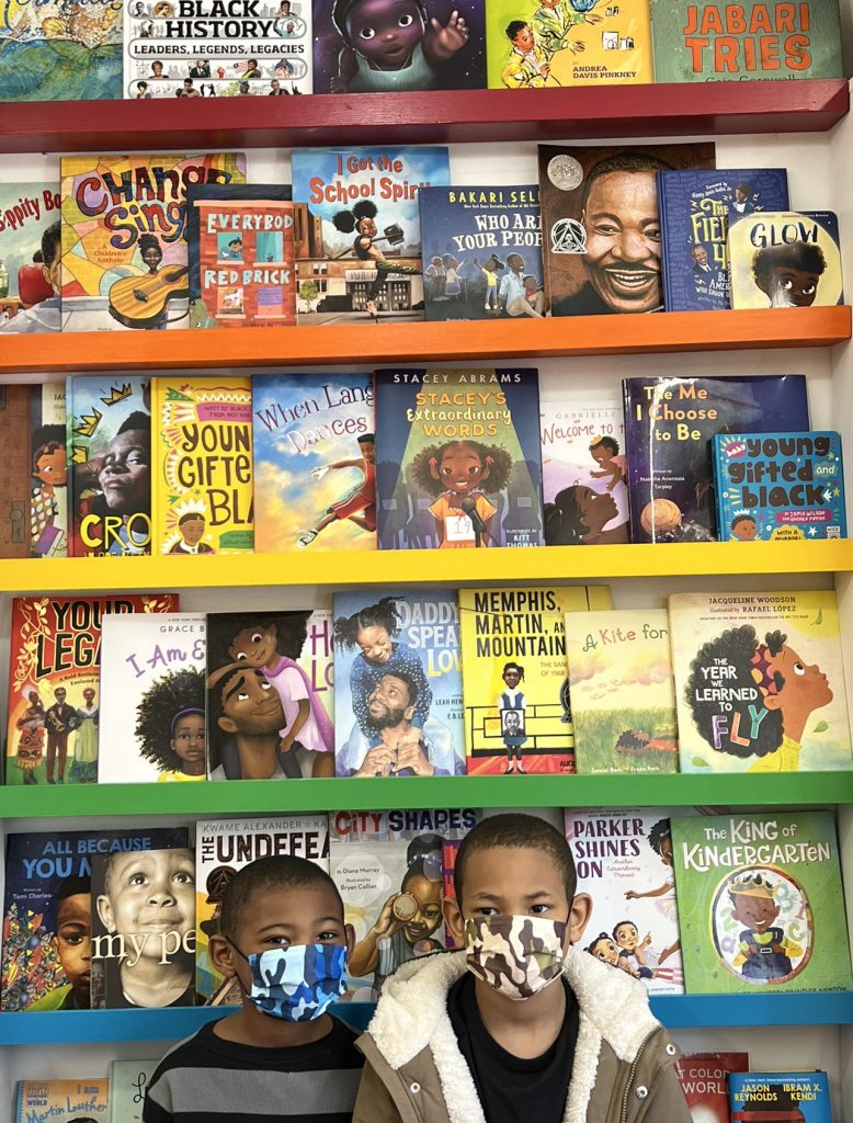 Boys wearing masks stand in front of shelves displaying books with diverse black characters